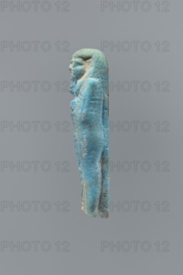 Ushabti, mummy-shaped figurine, back supported on a flat pillar. Feet missing. Arms crossed on the chest, right folded over left. Hoes in both hands. In the right hand, a string on which a basket is slung over the left shoulder. Slightly convex tools. On the head, a smooth, three-part wig. Round face, clearly marked large eyes, wide nose, mouth in a slight grimace. Engraved inscription, placed in one column on the front of the legs. Text illegible., 404–343 BC; Late Period, 28th–30th Dynasty, Egypt. Height 7 cm., A small vertical figurine rendered in shades of blue and turquoise with areas of greenish patina and small patches of beige and brown, shown in profile against a neutral gray background, depicting a standing human-like figure with a headdress and a braided or lobed hairstyle, arms held close to the torso with hands crossed over the chest, a smooth elongated body and subtle surface texture with visible pitting and wear.
