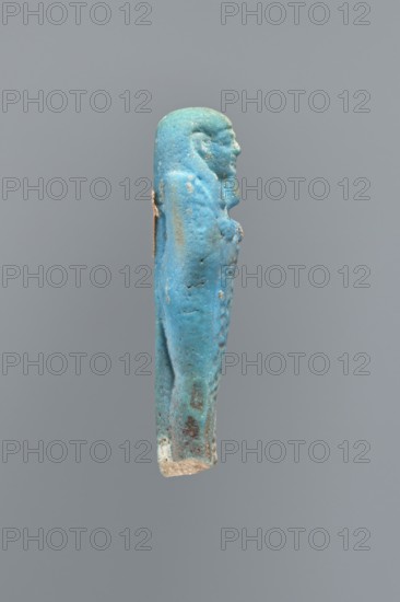 Ushabti, Mummy-shaped figurine, back supported on a flat pillar. Feet missing. Arms crossed on the chest, right folded over left. Hoes in both hands. In the right hand, a string on which a basket is slung over the left shoulder. Slightly convex tools. On the head, a smooth, three-part wig. Round face, clearly marked large eyes, wide nose, mouth in a slight grimace. Engraved inscription, placed in one column on the front of the legs; text illegible. 404–343 BC; Late Period, 28th–30th Dynasty, Egypt. Ushabti; height 7 cm., A small sculpted figure viewed in profile against a plain gray background, predominantly turquoise blue with areas of lighter blue and pale green patina, showing a human form with a rounded head, facial features, and a long body wrapped or clothed; there are a few rougher, weathered spots of brown and beige, and a small patch of white near the lower end, with some subtle darker blue and gray speckling across the surface.