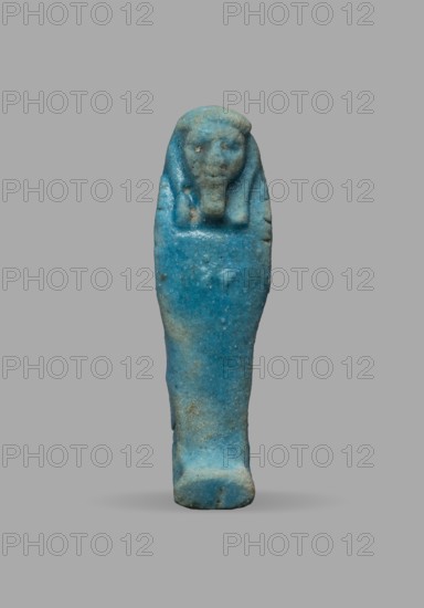 Ushabti, Wigs, funerary figurine, grave goods, mummy-shaped figurine. The arms are not distinct, with a slight convexity of the clenched hands. Delicate outline of tools on the arms. On the head is a smooth, tripartite wig. The face is large, round, with large eyes, a wide nose, and a slightly grimacing mouth. Not inscribed. Date: 525–332 BC, Late Period, 27th–30th Dynasty, Egypt. Ushabti; height 5.1 cm, width 1.5 cm., A small statuette shaped like a wrapped figure stands against a plain background, predominantly blue with areas of lighter and darker blue and small patches of brownish discoloration; the figure has a rounded head with simple facial features and a headdress-like form, a tapered body resembling a shroud, and a slightly wider base, with a rough textured surface and a few small pits and irregularities.