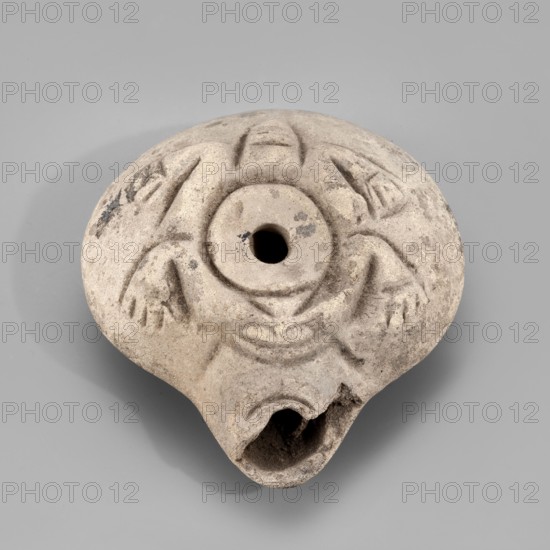 Lamp, Oil lamp with a circular container and a slightly forward burner. Decorated on the top with a schematic image of a frog surrounding the filler hole. The letter A is carved on the bottom. 3rd–4th century, Egypt. Oil lamp; height 3.4 cm, width 8.1 cm, depth 7.3 cm., A round, worn object with a central circular hole and a broken, curved opening at one edge, made of a light gray-beige material with darker gray spots and patches; its surface shows carved, raised decorative motifs forming curved lines and triangular shapes around the center, and the object casts a soft shadow on a smooth light gray background.