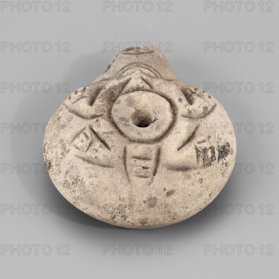 Lamp, Oil lamp with a circular container and a slightly forward burner. Decorated on the top with a schematic image of a frog surrounding the filler hole. The letter A is carved on the bottom. 3rd–4th century, Egypt. Oil lamp; height 3.4 cm, width 8.1 cm, depth 7.3 cm., A rounded object with a central round opening and a short protruding spout, made of a rough, light-colored material showing areas of darker discoloration and spots, with shallow carved linear and geometric grooves radiating from the center and subtle raised ridges around the opening; the surface displays a few smudges and darker marks, and the object rests on a flat, smooth gray background.