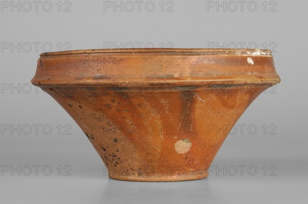 Bowl, Ancient Black Sea region, end of 1st–beginning of 2nd century AD, Crimea, vessel, height 6.1 cm, width 11.2 cm, A low, wide shallow bowl with a flared profile sits on a smooth surface; the vessel shows an earthy, terracotta orange color with darker brown and black spotting and areas of wear, along with a few paler, almost beige patches where the surface is abraded. The rim is slightly raised and has a similar orange-brown tone with small chips and rough edges. The body of the bowl has a matte, textured finish with scratches and irregular discoloration, and the base is narrower and slightly darker than the upper portion. The background is a uniform light gray.
