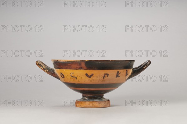 Black-figure kylix with Greek inscription, Archaic kylix form between a band/ribbon kylix and an eye kylix, 540–520 BC, Attica, kylix vessel, height 16.2 cm, width 22.2 cm, A shallow, two-handled ceramic bowl sits on a rounded foot against a neutral gray background, showing a warm ochre body with broad black bands near the rim and base; the handles and upper rim are painted dark, with some worn areas revealing lighter clay beneath, and there are several small dark brushmarks along the ochre band; overall colors visible include ochre, black, brown, and hints of beige from worn spots.