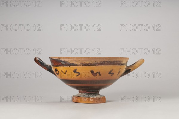 Black-figure kylix with Greek inscription, Archaic kylix a form between a band kylix and an eye kylix, 540–520 BC, Attica, kylix vessel, height 16.2 cm, width 22.2 cm, A shallow ceramic bowl with a rounded foot and two horizontal handles sits centered against a light gray background, showing tones of warm orange, dark brown, black, and lighter beige; the bowl's exterior has a broad orange band with a darker band above and below, dark brush marks decorating the orange area, and areas of worn black along the rim and handles, with the foot showing patches of lighter wear and some grayish residue.