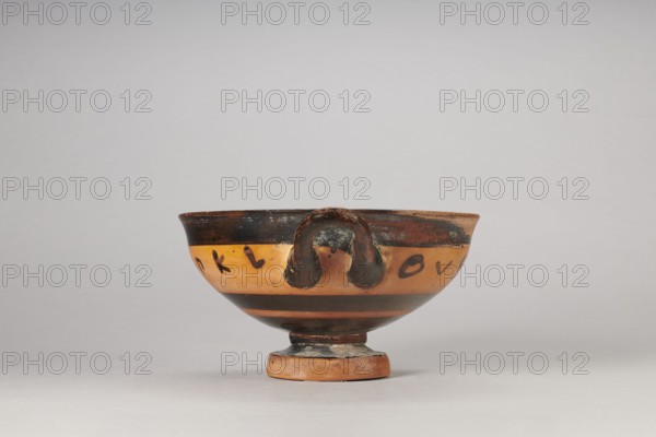 Black-figure kylix with Greek inscription, Archaic kylix form between a band/ribbon kylix and an eye kylix, 540–520 BC, Attica, kylix vessel, height 16.2 cm, width 22.2 cm, A small ceramic bowl with a rounded body and a short pedestal base sits centered against a pale gray background; the bowl shows a banded decoration with a dark rim and dark horizontal stripes contrasting against a warm orange-tan field, and a pair of small looped handles with darkened surfaces projecting from the rim; the surface displays areas of wear and darker mottling, with visible variation in the orange, brown, and black tones across the vessel.