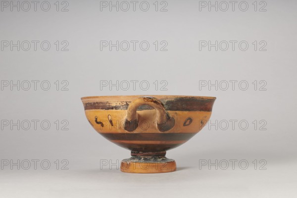 Black-figure kylix with Greek inscription, Archaic kylix a form between a ribband kylix and an eye kylix, 540–520 BC, Attica, kylix vessel, height 16.2 cm, width 22.2 cm, A shallow ceramic bowl on a small circular foot, shown from the front, with a single rounded handle attached to the rim and body; the surface shows bands and decorative marks in shades of orange, tan, brown, and black, with areas of wear and darker patina around the handle and rim, all set against a neutral pale gray background.