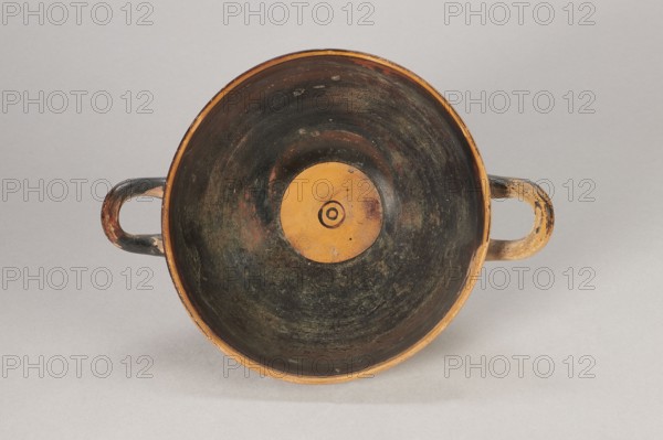 Black-figure kylix with Greek inscription, Archaic kylix form between a band/ribbon kylix and an eye kylix, 540–520 BC, Attica, kylix vessel, height 16.2 cm, width 22.2 cm, A top-down view of a shallow round bowl with two loop handles on opposite sides, showing interior surfaces that are predominantly dark with worn areas revealing brownish and ochre tones, a central circular ochre area with concentric rings, handles that are mottled with dark brown, black, and ochre, and a neutral light gray background.
