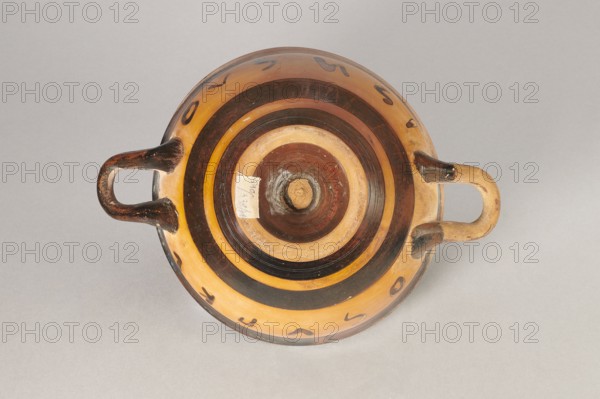 Black-figure kylix with Greek inscription, Archaic kylix form between a ribbon kylix and an eye kylix, 540–520 BC, Attica, kylix vessel, height 16.2 cm, width 22.2 cm, A round shallow ceramic bowl with two looped handles sits on a neutral gray background; the bowl shows concentric circular bands in shades of brown and tan with darker brown rings and lighter tan rings, and a few small dark painted marks around the rim; the handles are darker brown with areas of lighter tan where the glaze is worn, and the center of the bowl has a slightly raised circular hub with mixed brown and tan tones.