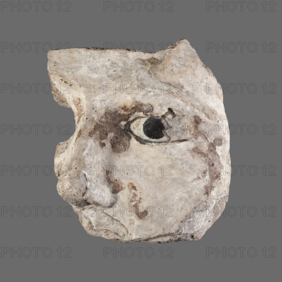 Stucco mask. Fragment of a sarcophagus mask left cheek, nose and left eye inlaid with white and black glass. Sarcophagus mask; masks, faces. Roman period, 2nd century; Egypt. Height 9.7 cm; width 11 cm., A sculpted fragment of a face shown in profile against a gray background, carved from a pale stone with an uneven, weathered surface; the visible portion includes a prominent nose, an eye with a dark circular center surrounded by lighter tones, and the suggestion of a mouth and cheekbone, with patches of darker brown and tan discoloration and rough texture across the pale off-white surface, and some subtle gray and beige variations in the stone.