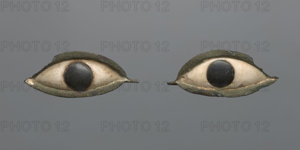 Pair of eyes from a sarcophagus mask. A pair of eyes probably originally placed in a sarcophagus mask. The whites are made of white stone, the iris of black stone. The whole is set in a copper setting. The copper setting is chipped at the edges, with larger losses on the outer side of the eyes. c. 1069–332 BC; Third Intermediate Period–Late Period, Egypt. Sarcophagus mask. Length: 6 cm part a, 6 cm part b., A pair of eye-shaped objects set against a gray background, each with an off-white oval center and a dark circular disk near the middle, framed by textured outer rims that appear greenish and brown with areas of rough, weathered surface; the off-white centers show subtle beige and tan discoloration and the dark disks have worn lighter scratches, while the surrounding rims display variations of green, brown, and hints of bluish corrosion.