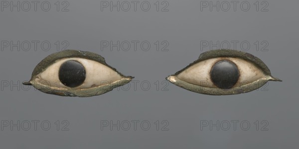 Pair of eyes from a sarcophagus mask. A pair of eyes probably originally placed in a sarcophagus mask. The whites are made of white stone, the irises of black stone. The whole is set in a copper setting; the copper setting is chipped at the edges, with larger losses on the outer side of the eyes. c. 1069–332 BC Third Intermediate Period–Late Period, Egypt. Sarcophagus mask. Length: 6 cm part a, 6 cm part b., A pair of symmetrical, eye-shaped objects set against a solid gray background, each composed of an off-white, curved central form resembling an eye sclera with a dark circular element at its center, surrounded by a textured, irregular outer rim that appears greenish with spots of brown and darker patina; the off-white areas show subtle brownish discoloration and surface marks, while the dark central circles are glossy and black with some light scratches.