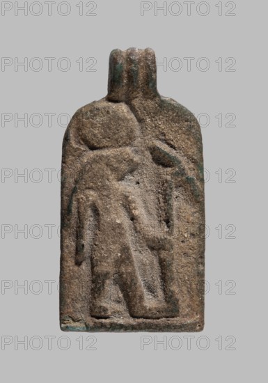 Amulet plaque with the figure of Re: plaque with a flat relief figure of a falcon-headed deity with a sun disk on his head, turned to the right and holding a uas scepter; pendant at the top. Re mythology. 7th–4th century BC, Late Period, Egypt. Amulet. Height 2.6 cm; width 1.4 cm., A rectangular, vertically oriented carved stone or clay plaque with rounded top and a small protruding neck, showing a low-relief depiction of a walking human-like figure; the surface is rough and weathered with visible pitting and grain, and there are traces of greenish discoloration and darker brown areas against an overall tan to light brown base color, set on a plain light gray background.
