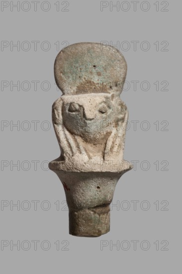 Amulet - falcon head on a column; papyrus capital of the column shows a falcon head in a striped wig topped with a disc, with a pendant at the back. 332–30 BC, Ptolemaic Period, Egypt. Amulet; height 3.6 cm, width 1.5 cm., A small sculpted object resembling a stylized crouching figure with a rounded top, rough textured surface, and simplified facial features; the piece shows a head with a flat forehead area above recessed eyes and a suggestion of limbs folded close to the body, all carved in a single piece that tapers into a cylindrical base. The colors visible are pale beige, light tan, muted green, and small areas of brown and gray, with subtle mottling and weathered patches across the surface.