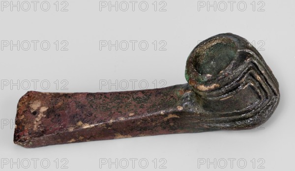 Pounder, unknown Near Eastern workshop; pounder with a low, ribbed, cylindrical socket and a blade in the form of a massive wedge set at a slight angle to the shaft. 1st half of the 2nd millennium BC; Middle Bronze Period. Western Iran Iran. Weapon. Geometric ornaments; edged weapon; material culture; Eastern Gaulish scenography. Height 4.6 cm, width 12.5 cm, depth 3.2 cm, weight 324 g., An irregularly shaped metallic object with a long rectangular shaft transitioning into a rounded cup-like end; the surface shows pitting and rough texture with areas of corrosion. The colors visible include dark brown, reddish-brown, green, black, and patches of lighter beige or tan. The cup end has layered, ridged detailing and a hollow interior that appears darker green and black, while the shaft displays mottled patches of reddish-brown and darker speckling with occasional lighter spots.