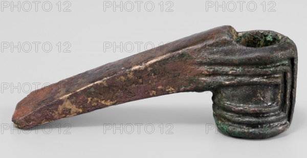 Pounder, unknown Near Eastern workshop; pounder with a low, ribbed, cylindrical socket and a blade in the form of a massive wedge set at a slight angle to the shaft. 1st half of the 2nd millennium BC; Middle Bronze Period. Western Iran findspot: Iran. Weapon. Material culture: geometric ornaments, edged weapons. Height 4.6 cm; width 12.5 cm; depth 3.2 cm; weight 324 g., A close-up of a weathered metal object with a tapered, wedge-like extension attached to a rounded, hollow end; the surface shows rough, pitted textures and layered ridges on the rounded portion. The visible colors include brown, green, dark gray, and patches of goldish tones, with mottled areas where the colors blend and a few lighter highlights on raised edges. The object rests on a neutral light-gray background and casts a soft shadow beneath it.