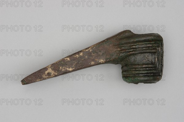 Pounder, unknown Near Eastern workshop; pounder with a low, ribbed, cylindrical socket and a blade in the form of a massive wedge set at a slight angle to the shaft. 1st half of the 2nd millennium BC Middle Bronze Period. Western Iran, Iran. Weapon. Geometric ornaments; edged weapon. Height 4.6 cm, width 12.5 cm, depth 3.2 cm, weight 324 g., An aged metal object lies against a pale gray background, showing a long tapered shaft that broadens into a rectangular block with rounded edges; the surface displays a mottled mix of dark brown, green, and patches of lighter tan or beige, with visible textured corrosion and layered ridges on the block end.