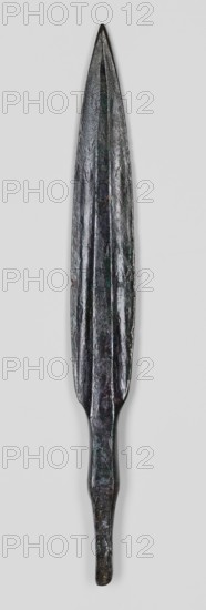 Javelin head, unknown Near Eastern workshop, weapon; flat spear head with an elongated lanceolate blade with a high rib of rectangular cross-section, transitioning at the base into a short, rectangular socket, which transitions into an equally short, tapering, lath-like spike of rectangular cross-section. Luristan bronzes, militaria, ranged weapon, material culture, Eastern Gaulish Scenography. 9th–6th century BC end of the 2nd millennium–first half of the 1st millennium BC; Iron Period. Near East; Luristan. Height 15.3 cm, width 1.8 cm, depth 0.7 cm., An elongated metal object with a pointed tip and a tapered handle, showing an overall dark metallic color with areas of greenish and brownish patina, a central raised ridge running along its length, a textured and slightly irregular surface, and a narrow rounded end opposite the tip, positioned against a pale gray background.