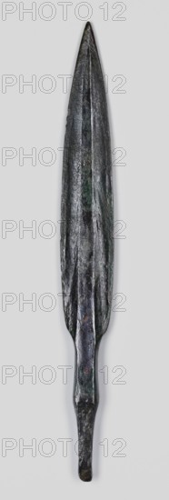 Javelin head, unknown Near Eastern workshop; flat spear head with an elongated lanceolate blade with a high rib of rectangular cross-section, transitioning at the base into a short rectangular socket, which transitions into an equally short, tapering lath-like spike of rectangular cross-section. Material: bronze Luristan bronzes?. Function: weapon, ranged weapon/militaria. Style/period: Iron Age, 9th–6th century BC end of 2nd millennium–first half of 1st millennium BC. Place of origin/find: Luristan, Near East. Dimensions: height 15.3 cm; width 1.8 cm; depth 0.7 cm., A long, narrow metal object with a pointed tip and a tapered base, shown vertically against a light gray background; the surface appears rough and uneven with visible ridges and a central raised ridge running along much of its length; the color is predominantly dark gray to black with areas of greenish patina and small patches of brownish discoloration; the lower section narrows into a thinner, slightly flattened shaft bef...