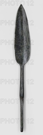 Javelin head, unknown Near Eastern workshop. Flat spearhead with a lanceolate leaf, with a poorly defined rib of rectangular cross-section, passing at the base into a round socket, which passes into an equally long, tapering, lath-like spike of rectangular cross-section. Possibly Luristan, Near East; 9th–6th century BC end of the 2nd millennium–first half of the 1st millennium BC; Iron Period. Weapon ranged weapon, militaria, material: bronze Luristan bronzes?. Dimensions: height 17.4 cm; width 2.3 cm; depth 0.7 cm., A single elongated metal object with a pointed, leaf-shaped blade that tapers into a narrow cylindrical shaft; the surface of the blade and shaft appears textured and slightly irregular with visible wear. The object shows dark colors including black and deep green with subtle variations and mottling, and it rests against a plain light gray background.