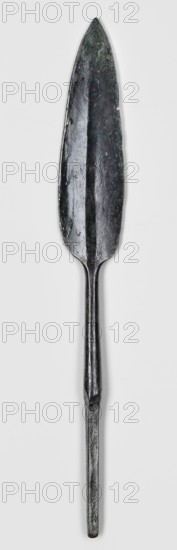 Javelin head, unknown Near Eastern workshop. Flat spearhead with a lanceolate leaf, with a poorly defined rib of rectangular cross-section, passing at the base into a round socket, which passes into an equally long, tapering, lath-like spike of rectangular cross-section. Weapon; militaria; ranged weapon; material culture. 9th–6th century BC end of the 2nd millennium–first half of the 1st millennium BC; Iron Period. Near East; Luristan. Height 17.4 cm, width 2.3 cm, depth 0.7 cm., A long metallic spearhead with a pointed leaf-shaped blade and a narrow socketed shaft, showing a dark greenish-black patina with areas of lighter green and brownish discoloration, visible surface pitting and rough texture, a central shallow ridge running along the blade, and the shaft tapering into a cylindrical tang against a plain light gray background.