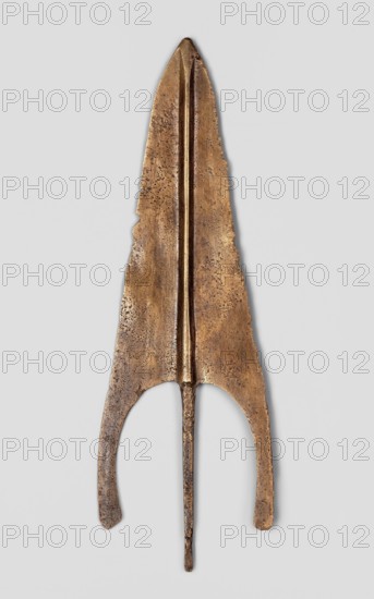 Javelin head, unknown Near Eastern workshop; weapon, flat spearhead with a long sagittal blade with a high rib of circular cross-section transitioning at the base into a tapering, lath-like spike of rectangular cross-section; material: bronze Luristan bronzes ?; function: ranged weapon/militaria; style/period: Iron Age, 9th–6th century BC end of the 2nd–first half of the 1st millennium BC; place of origin/find: Luristan, Near East; dimensions: height 18.1 cm, width 5.9 cm, depth 0.6 cm., An elongated metallic object with a pointed top and a tapering shaft, showing a warm bronze to brown patina with areas of darker brown and mottled texture, with a central raised ridge running lengthwise, irregular edges and a rounded cutout near the lower portion forming two curved prongs, set against a pale gray background.