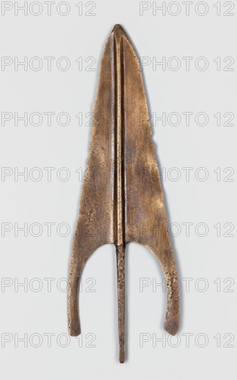 Javelin head, unknown Near Eastern workshop; weapon, Luristan bronzes, militaria, ranged weapon, material culture, Eastern Gaulish Scenography. Flat spearhead with a long, sagittal blade with a high rib of circular cross-section, transitioning at the base into a tapering, lath-like spike of rectangular cross-section. 9th–6th century BC end of the 2nd–first half of the 1st millennium BC; Iron Period. Near East; Luristan. Height 18.1 cm; width 5.9 cm; depth 0.6 cm., A long, narrow metallic object with a tapered, pointed top and a pair of curved extensions near the bottom, displayed against a pale background; the object has a central raised ridge running vertically and a thin shaft projecting downward from the center; the metal surface shows a worn, pitted texture and uneven edges; visible colors include brown, bronze, golden, and darker brown tones.