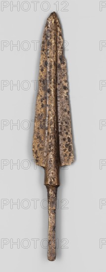 Javelin head, unknown Near Eastern workshop. Luristan bronzes, militaria, ranged weapon, Eastern Gaulish Scen. Flat spearhead with an elongated triangular blade with a high rib of circular cross-section, transitioning at the base into a short sleeve of circular cross-section, which transitions into a long, tapering, lath-like spike of rectangular cross-section. 9th–6th century BC; Iron Period end of the 2nd millennium–first half of the 1st millennium BC. Near East; Ancient Near East. Weapon. Height 12.5 cm, width 2.1 cm, depth 0.8 cm., An elongated metallic object with a tapered, leaf-shaped upper section and a narrow cylindrical lower shaft, showing a rough, pitted surface texture. The upper section has a central raised ridge and flared edges with uneven, jagged contours. The metal displays mottled colors including brownish gold, dark brown, and areas of dull gray, with darker irregular spots scattered across the surface. The lower shaft is narrower and shows similar roughness and col...