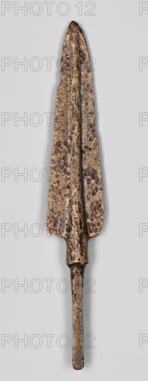 Javelin head, unknown Near Eastern workshop; Luristan bronzes, militaria, ranged weapon; Flat spearhead with an elongated triangular blade with a high rib of circular cross-section, transitioning at the base into a short sleeve of circular cross-section, which transitions into a long, tapering, lath-like spike of rectangular cross-section. 9th–6th century BC; Iron Period end of the 2nd millennium–first half of the 1st millennium BC. Near East. Weapon. Height 12.5 cm, width 2.1 cm, depth 0.8 cm., A weathered metallic spearhead with a long tapered blade and a narrow tang extending downward, showing a rough pitted surface and irregular edges; the object displays a mix of brown, dark brown, and golden tones with darker spots and patches, set against a light gray background.