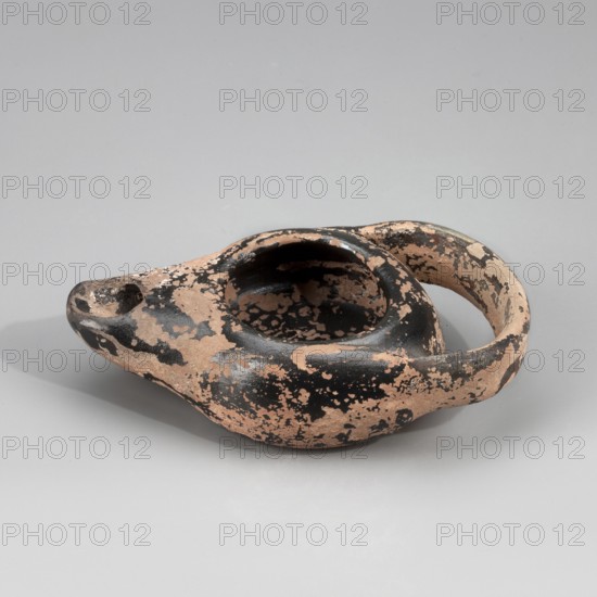 Black-finished lamp, unknown Attic workshop. Lamp made on a wheel. Circular, flattened vessel with curved vertical walls. Flat, circular base. Vertically attached, ribbon-like handle. Arms sloping downward, outward. Large oil hole disc missing. Medium-length burner, slightly upturned. Entire piece coated with black varnish engobe/firnis before firing. Attic clay. 425–400 BC, Myrmekion; Attica origin. Oil lamp; height 2.5 cm, width 5.6 cm, length 9.6 cm., An aged ceramic oil lamp with a rounded body, a raised circular reservoir, a tapered spout, and a curved handle, showing a mottled surface with patches of black and pale terracotta, placed on a smooth light gray background.