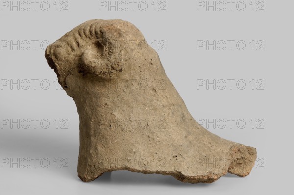 Fragment of a ram figurine preserved from half the snout to the neck and a small piece of the back. The snout is impressed in a mold with lens-shaped eyes surrounded by thick eyelids and several ridges above the eyes, converging in the center of the snout. The ram's horns, curled around the ears, are only marked by indentations and emerge from the top of the animal's head and are provided with transverse indentations imitating the natural texture of ram horns. Rusty clay, coarse-grained, crumbling. 1st half of the 6th century BC, Etruria. Antefix fragment. Height 13 cm, width 15 cm., A roughly textured object with a weathered, stone-like surface in shades of beige and light brown, showing a rounded top with a spiral-shaped protrusion and a slope that widens toward the base; the surface has small pits and granular irregularities and edges that appear chipped, set against a plain light gray background.