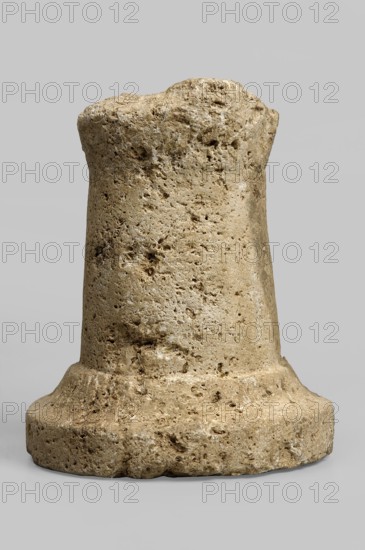Thymiaterion, missing the upper part of the hearth and approximately half of the base. Thymiaterion with a cylindrical, smooth shaft, slightly tapering towards the top, provided with a high half the height of the shaft profiled base. The base consists of two elements — a vertical lower part and a slightly concave upper part, on which stands the thymiaterion's head, slightly spaced from the edge. Late Hellenistic period, Etruria, varia; height 13.3 cm, width 11 cm., A roughly cylindrical stone object with an irregular, pitted surface sits against a plain light gray background; the stone shows a range of beige, tan, and light brown tones with some darker brown speckles and small cavities, a wider rounded base tapering slightly toward a blunt, uneven top, and visible rough texture and porous holes across its surface.