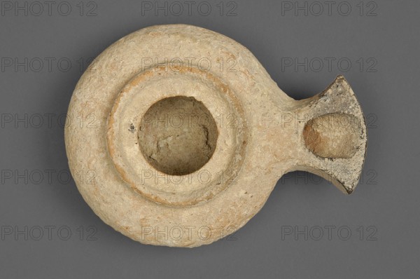 Herodian oil lamp, a small clay lamp with a slightly flattened circular container and a fan-shaped burner. The large round filling opening is surrounded by a convex ring approximately 0.5 cm larger than the opening. Orange clay with light cream engobe. 1st century, Palestine, oil lamp., A round, shallow vessel with a central circular basin and an extended spout-like projection, made of rough, worn material showing a sandy beige and light tan surface with small patches of darker brown and faint orange tones; the rim around the basin is slightly raised, and the basin interior is textured and similar in color to the exterior; the spout area has a darker, weathered edge with mottled gray and brown discoloration; the background is a uniform medium gray.