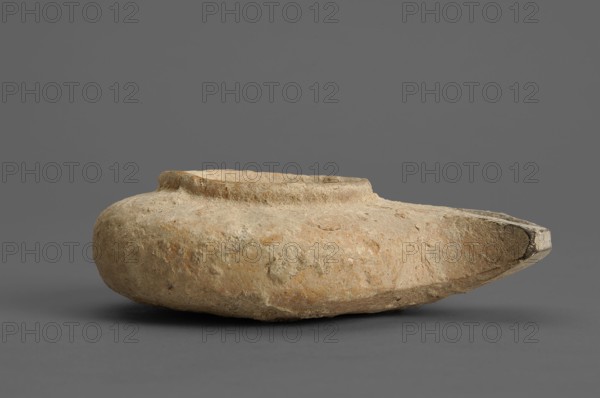 Herodian oil lamp, small clay lamp with a slightly flattened circular container and a fan-shaped burner. Large round filling opening surrounded by a convex ring about 0.5 cm larger than the opening. Orange clay with light cream engobe. 1st century, Palestine. Oil lamp., A roughly disc-shaped object with a shallow, slightly raised rim sits against a plain background, showing a textured, weathered surface with irregular pitting and worn edges; the object displays earthy tones of beige, tan, and light brown with hints of pale ochre and small darker spots, and the background and surface beneath the object are shades of gray.