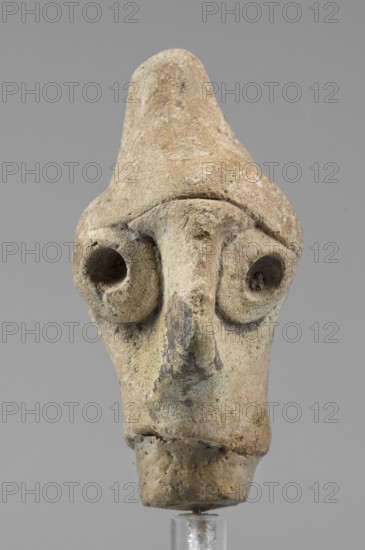 Idol head, unknown Syrian workshop, terracotta, idols, Northwestern Syria, male head in a conical headdress rounded at the tip. Flattened, triangular nose projecting forward. Eyes and ears preserved as glued tablets with a drilled hole in the center. A narrow strip of clay glued above the forehead. 1st half of the 2nd millennium BC, Orontes Region, figurine, height 4.5 cm, width 2.5 cm., A weathered sculpted head made of a beige to light brown material stands against a plain gray background, showing a conical top, deep rounded eye sockets with hollow centers, a prominent elongated nose with darker grayish-brown discoloration, and a squared lower portion that tapers down to a rough cylindrical base; the surface has patches of darker brown and gray, small pits and rough texture, and a few areas of slight whitish residue.
