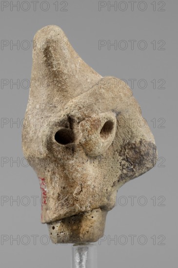 Idol head, unknown Syrian workshop, terracotta, idols, Northwestern Syria. Male head in a conical headdress, rounded at the tip. Flattened, triangular nose, protruding forward. Eyes and ears trace preserved made of glued tablets with a hole drilled in the center. A narrow strip of clay is glued above the forehead. 1st half of the 2nd millennium BC, Orontes Region. Figurine, height 4.5 cm, width 2.5 cm., A weathered beige and tan stone object with a rough, pitted surface sits against a plain gray background; its irregular form includes a prominent rounded projection at the top, a pair of hollowed circular cavities on one side, and a narrower base with small perforations, with areas of darker brown and faint reddish discoloration visible on parts of the surface.