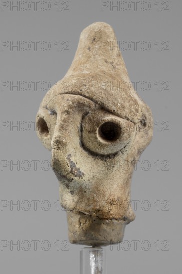 Idol head, unknown Syrian workshop, terracotta, idols, Northwestern Syria. Male head in a conical headdress, rounded at the tip. Flattened, triangular nose, protruding forward. Eyes and ears traces preserved made of glued tablets with a hole drilled in the center. A narrow strip of clay is glued above the forehead. 1st half of the 2nd millennium BC, Orontes Region. Figurine, height 4.5 cm, width 2.5 cm., A close-up of a small sculpted head made of a rough, beige-brown material with weathered texture and irregular surface spots; the head has a pointed top, deep rounded eye sockets set into a pronounced brow ridge, and a long nose extending down toward a small mouth area; the surface shows darker brown and grayish discolorations and patches of lighter beige; the object is mounted on a clear stand against a plain gray background.