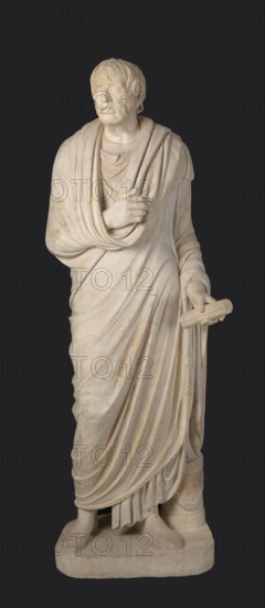 Depiction in the Pseudo-Seneca type, identified as a portrait of Aristophanes or Hesiod; maker, pseudo-Seneca, male portrait in Roman sculpture; togate figure; end of the 1st century BC previously dated 4th quarter of the 1st century BC; Latium; sculpture; weight 610 kg; height 183 cm; width 70 cm; depth 55 cm; base width 62.5 cm; base depth 47 cm., A marble statue of a robed standing man against a dark background, with light beige to off-white stone showing subtle veins and darker weathered spots; the figure has short textured hair and a beard, a solemn facial expression with closed or lightly-open eyes, and a draped cloak wrapped over one shoulder and across the chest with flowing folds and layered fabric; one hand rests near the chest holding part of the drapery while the other extends slightly forward holding a cylindrical object; the lower robes form deep vertical folds that fall to the base, where the statue stands on a rectangular plinth with visible feet and sandals.