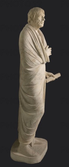 Depiction in the Pseudo-Seneca type, currently identified as a portrait of Aristophanes or Hesiod; creator, pseudo-Seneca, male portrait, Roman sculpture; marble; toga; end of the 1st century BC previously dated to 4th quarter of the 1st century BC; Latium; sculpture; weight 610 kg; height 183 cm; width 70 cm; depth 55 cm; base width 62.5 cm; base depth 47 cm., A full-length pale stone statue of a robed standing man shown in profile against a dark background, with a textured face and short wavy hair, a beard, and a contemplative expression; he is draped in layered flowing garments with visible folds and creases, one arm crossed near his chest and the other extended holding a rolled object; he stands on a rectangular base wearing footwear that is partly visible; the visible colors are pale off-white and beige of the stone, with a dark gray background.