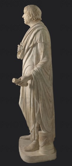 Depiction in the Pseudo-Seneca type, identified as a portrait of Aristophanes or Hesiod; creator, pseudo-Seneca male portrait in Roman sculpture style; toga; end of the 1st century BC previously dated 4th quarter of the 1st century BC; origin: Latium; sculpture; weight 610 kg; height 183 cm; width 70 cm; depth 55 cm; base width 62.5 cm; base depth 47 cm., A carved pale stone statue of a standing robed male figure shown in profile against a dark background, with short wavy hair and a beard, wearing layered drapery that falls in flowing folds over his shoulders and down his body, one hand holding the edge of the robe near his chest and the other extended forward holding a rolled scroll, the garment forming soft curved lines and pleats over the arms and legs, the feet shod and resting on a rectangular base accompanied by a cylindrical object beside one foot, the stone surface showing subtle variations of off-white, cream, and light beige tones.