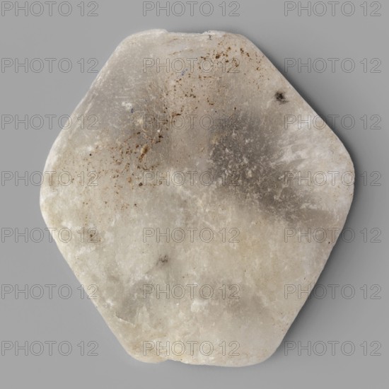 Rhomboid bead, unknown Northern Mesopotamian workshop. Rhomboid bead with smooth surfaces, with two corners cut off at the ends of the axis, along which a hole for stringing was drilled. 1st half of the 3rd millennium BC; Early Jazira III period Early Dynastic III; ca. 2600–ca. 2350 BC. Northern Mesopotamia Khabur basin, Tell Rad Shakra northeastern Syria. Found in children's graves. Decoration. Height 2.2 cm, width 2.08 cm, depth 0.8 cm., A roughly hexagonal stone with rounded edges resting on a gray background, showing a mix of off-white, beige, light gray, and darker gray areas, with a few scattered brownish specks and subtle translucent patches across its surface.
