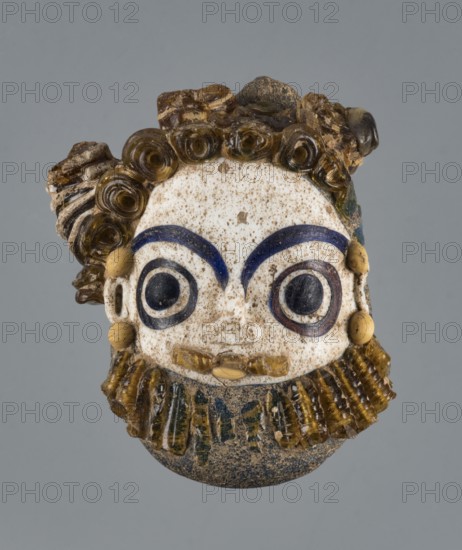 Apotropaic pendant in the shape of a male head; bearded head pendant; Carthage Tunisia; technique on core; Pendant in the form of a bearded man's head; 4th–3rd century BC; jewelry; amulet; height 8 cm, width 6.4 cm, depth 4 cm., A small sculpted face with a pale, speckled surface framed by coiled and ridged brown hair and a matching brown beard, featuring prominent curved dark blue eyebrows, large circular dark eyes set within rings of blue and brown, rounded beige ear ornaments, a small beige mouth, and a mottled dark base beneath the beard, all set against a plain gray background.