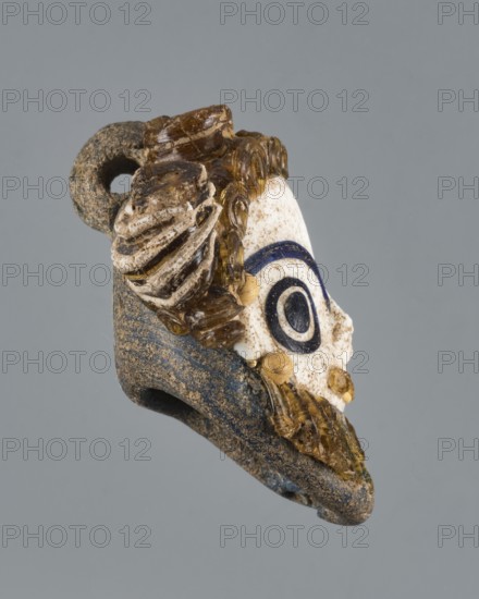 Apotropaic pendant in the shape of a male head; bearded head pendant; Carthage Tunisia; technique on core; pendant in the form of a bearded man's head; 4th–3rd century BC; jewelry amulet; height 8 cm, width 6.4 cm, depth 4 cm., A small carved object photographed against a gray background showing a profile view of a face-like form with a white surface and dark eye-shaped decoration, surrounded by textured brown and amber-colored elements that resemble stylized hair or curls, attached to a larger rough-textured base that is dark gray with sandy brown speckling; visible colors include gray, white, dark blue or black, brown, amber, and sandy tan.
