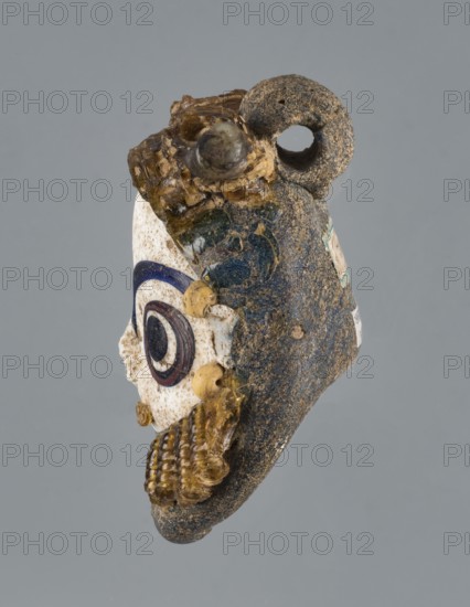 Apotropaic pendant in the shape of a male head, bearded head pendant, pendant in the form of a bearded man's head. 4th–3rd century BC, Carthage Tunisia. Technique on core. Jewelry amulet. Height 8 cm, width 6.4 cm, depth 4 cm., A worn object resembling a small sculpted head with a textured dark gray and brown surface, a loop at the top, and attached translucent yellowish glass elements; one side shows a white area with concentric circular decorations in dark blue and dark brown, and small round yellowish beads; the object displays a mix of colors including gray, brown, white, dark blue, dark brown, and yellowish amber, with rough and smooth textures and some glossy glassy parts.