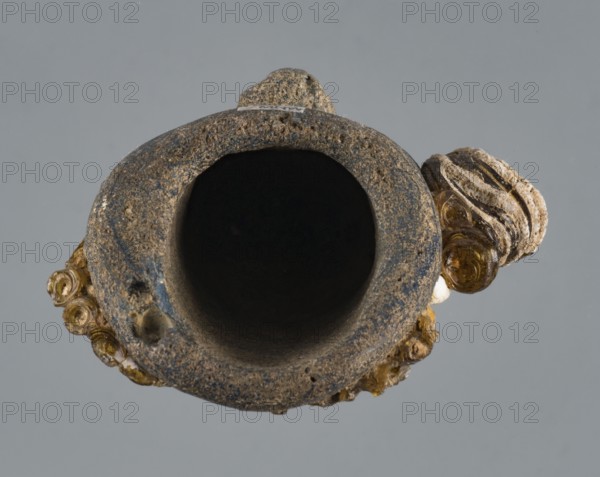 Apotropaic pendant in the shape of a male head; bearded head pendant; Carthage Tunisia; technique on core; pendant in the form of a bearded man's head; 4th–3rd century BC; jewelry amulet; height 8 cm, width 6.4 cm, depth 4 cm., A close-up view of a hollow cylindrical object with a rough textured surface in shades of brown and gray, showing an open dark interior; attached to its rim is a group of small spiral-shaped pieces in amber and translucent yellow tones, as well as some lighter beige and off-white layered elements; the background is a uniform muted gray.