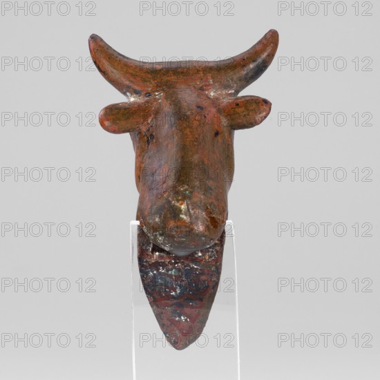 Stylized bull-headed protoma used as vessel decoration, typical Urartian cauldron ornament. Convex eyeballs are surrounded by a straight line; two horizontal lines mark the nostrils. Small protruding ears are plastically modeled; slightly curved, chipped horns are schematic. The neck is slightly elongated with a triangular bulge at the front. A rectangular cutout at the rear bottom served to mount the protoma on the rim of a cauldron. Material/function: protoma decoration. Date: 8th–7th century BC Urartu, ca. 850–585 BC. Place of manufacture: Urartu/Near East. Weight: 134 g., The image shows a brown sculptural object resembling a stylized animal head with upward-curving horns and small rounded protrusions on the sides, mounted on a clear support; the surface has mottled areas of darker brown, rusty red, and patches of greenish and whitish discoloration, with a smooth worn texture and subtle irregularities, set against a plain light gray background.