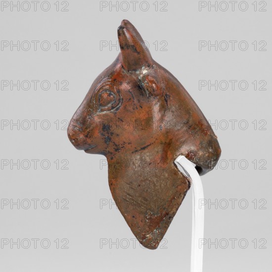 Bull-headed protoma, vessel decoration, unknown Urartian workshop; protoma, bull heads iconography, Eastern Gaulish Scenography. Stylized bull's head with linear details — large convex eyeballs surrounded by a straight line; two horizontal lines mark the nostrils. Small, protruding ears plastically modeled; schematically chipped horns slightly curved upward. Neck slightly elongated with a triangular bulge at the front. Rectangular cutout at the rear bottom for mounting the protoma on the rim of a cauldron. Typical Urartean cauldron decoration. 8th–7th century BC Urartu, ca. 850–585 BC. Near East; Urartu. Decoration. Weight 134 g., A small sculpted object shaped like an animal head, shown in profile against a light gray background, with a smooth, worn surface that is predominantly reddish-brown with areas of darker brown and hints of greenish-blue patina; the head has a pointed ear, a rounded snout, a carved circular eye detail, and a tapered lower portion, and it is mounted on a pale, ...