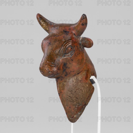 Bull-headed protoma, vessel decoration, unknown Urartian workshop. Protoma depicting a stylized bull's head with linear details: large convex eyeballs surrounded by a straight line, two horizontal lines marking the nostrils; small protruding ears plastically modeled; schematically chipped horns slightly curved upward. The neck is slightly elongated with a triangular bulge at the front. At the bottom rear is a rectangular cutout that served to mount the protoma on the rim of a cauldron. Typical Urartian art decoration of cauldrons. 8th–7th century BC Urartu, ca. 850–585 BC. Near East; manufactured in Urartu. Decoration; weight 134 g., A sculpted head of an animal with upward curved horns, a snout, and an ear visible on one side, shown against a plain light gray background; the surface shows a weathered patina in shades of reddish-brown, dark brown, and some greenish-blue spots, with areas of darker speckling and lighter worn patches; the object is mounted on a thin white support that is...