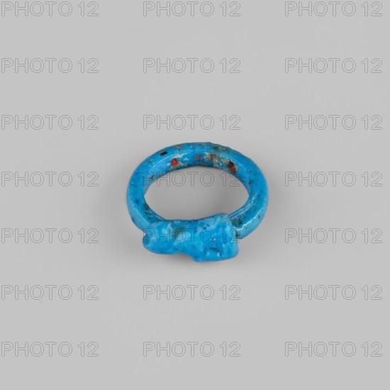 Ring with a kitten;, Sacred animal amulet ring, New Kingdom, 18th Dynasty, Egypt, ca. 1550–1295 BC. Materials and dimensions: ring/amulet, height 1.8 cm. Subject: cat/kitten., A small circular object with an irregular shape, predominantly bright blue with areas of darker blue and small specks of red and greenish-brown, sits centered on a smooth light gray background; the object has a rounded ring form with a slightly thicker, lumpy section on one side and a rough, pitted surface texture.