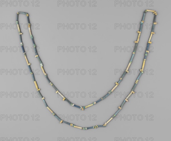 Necklace, disc-shaped and tubular beads; necklace consisting of 52 tubular beads, 118 flat beads and 21 double beads with 2 holes. Colors green and blue, individual beads yellowish. 664–332 BC; Late Period, Egypt. Jewelry; height 113 cm., A pair of beaded necklaces arranged in a loose double loop on a flat background, each necklace made of a sequence of small cylindrical and short tubular beads with occasional slightly larger spacer beads; the beads show a mix of muted blue, teal, green, beige, and pale yellow tones, with some beads appearing weathered or patinated, and the background is a smooth light gray.