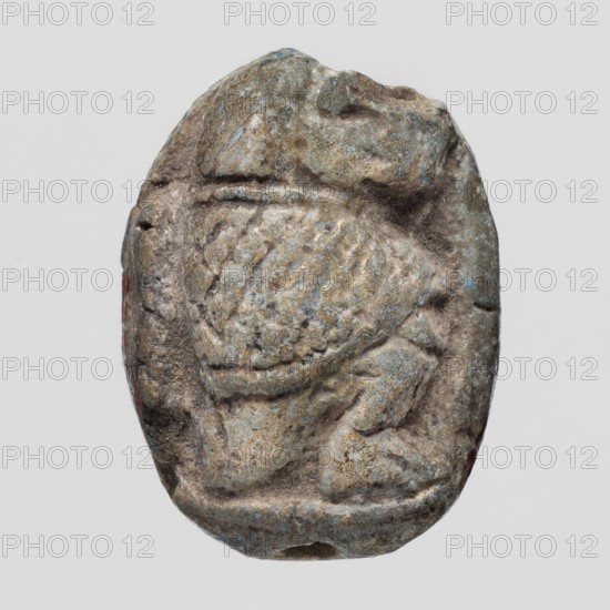 Scaraboid with a monkey. The upper surface of the scaraboid is decorated with a convex image of a seated baboon facing right, with the fur on its back marked with crisscrossing lines. On the base is depicted a lying sphinx lion? wearing a feathered crown, above it a solar disk. In front of the sphinx is the figure of a seated goddess Maat?, above the sphinx's back is visible a lying feather of Maat and possibly another symbol damaged. c. 1550–c. 1186 BC; New Kingdom, 18th–19th Dynasty, Egypt. Amulet, scaraboid; height 2.3 cm, width 1.7 cm., A small worn carved object with an oval outline showing a raised textured central area and surrounding indented grooves, exhibiting a rough surface with pitted and eroded details; the object displays a palette of muted colors including gray, brown, tan, and hints of bluish-green with tiny spots of reddish tone along the edges, and the background is a uniform light gray.